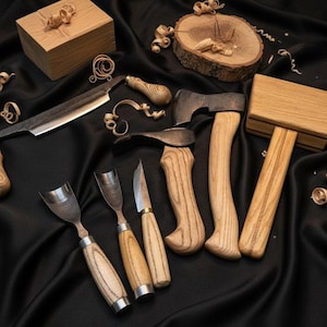 May include: A collection of woodworking tools, including a drawknife, gouges, a hatchet, and a mallet, arranged on a black satin surface. The tools have wooden handles and metal blades. Wood shavings are scattered around the tools.