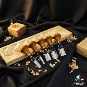 May include: A set of six wood carving chisels with wooden handles and silver blades, arranged on a carved wooden base. The chisels are displayed on a black satin surface, with wood shavings scattered around. A small wooden block and the American Handy Craft logo are also visible.