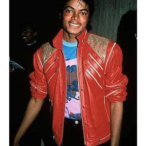 Puede incluir: Chaqueta de cuero roja con detalles dorados en los hombros y múltiples cremalleras. La chaqueta se lleva sobre una camiseta gráfica azul y rosa. La imagen muestra a una persona sonriendo.