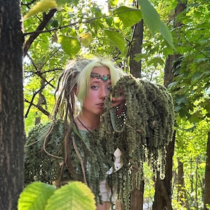 Puede incluir: Una persona con un top de ganchillo verde y un tocado a juego, adornado con un adorno decorativo para la cabeza. La persona est&aacute; en un entorno forestal, rodeada de exuberante follaje verde y luz solar. La est&eacute;tica general evoca un tema natural y terroso.