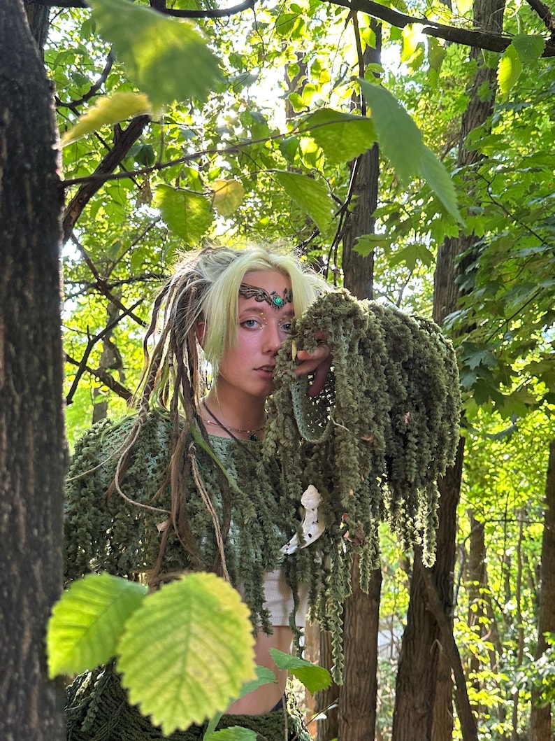 Puede incluir: Una persona con un top de ganchillo verde y un tocado a juego, adornado con un adorno decorativo para la cabeza. La persona est&aacute; en un entorno forestal, rodeada de exuberante follaje verde y luz solar. La est&eacute;tica general evoca un tema natural y terroso.