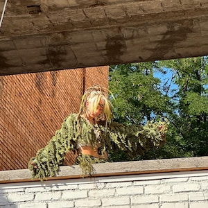 Puede incluir: Una persona con una prenda verde y frondosa con mangas largas y un tocado a juego. El atuendo se combina con un top y cintur&oacute;n met&aacute;licos. El fondo incluye una pared de ladrillo, una estructura de hormig&oacute;n y follaje verde.