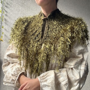 May include: A moss green shaggy knit capelet featuring a textured, fringed yarn construction and a single wooden toggle button closure at the neckline. The garment is worn over a long-sleeved, cream-colored blouse with a subtle floral pattern.