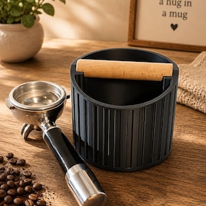 Könnte beinhalten: Ein schwarzer Abschlagbehälter für Espresso mit einem hellen Holzstab und einem passenden Siebträger mit schwarzem Griff und silbernem Metallsieb. Der Abschlagbehälter hat ein vertikal geripptes Außendesign und liegt neben Kaffeebohnen.