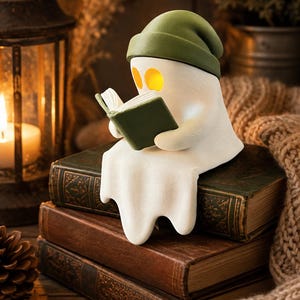 Könnte beinhalten: Eine weiße Geisterfigur mit einer dunkelgrünen Mütze, die auf einem Stapel aus drei alten Büchern sitzt und ein offenes, dunkelgrünes Buch hält. Der Geist hat leuchtend gelbe Augenhöhlen und sitzt auf dem obersten Buch des Stapels.