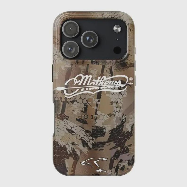 K&ouml;nnte beinhalten: Sch&uuml;tzende Handyh&uuml;lle mit Tarnmuster in Braun-, Beige- und Gr&uuml;nt&ouml;nen. Die H&uuml;lle zeigt den wei&szlig;en Text "Mathews" mit einem Pfeildesign. Die H&uuml;lle hat drei Kameralinsen und einen Blitz.
