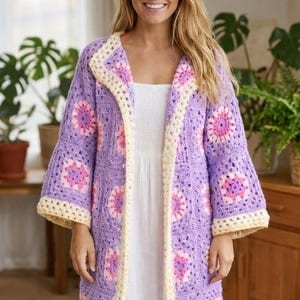 May include: A long, open-front crochet cardigan in a soft lavender hue. The cardigan features a floral pattern in pink and cream, with cream-colored trim along the edges and sleeves. The sleeves are flared, and the overall style is bohemian.