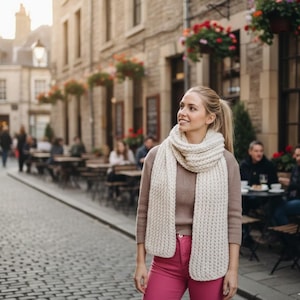 Könnte beinhalten: Eine Frau trägt einen cremefarbenen, grob gestrickten Schal und einen braunen Pullover, kombiniert mit einer rosa Hose. Der Hintergrund zeigt eine Kopfsteinpflasterstraße mit Außensitzplätzen in Cafés und Gebäuden mit Blumenampeln.