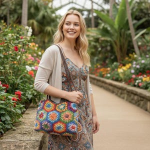 May include: A colorful crochet tote bag with a geometric floral pattern. The bag features a variety of colors, including blue, orange, yellow, and green. The bag is carried by a person wearing a patterned dress and a beige cardigan.