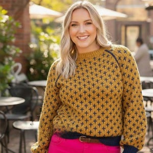 May include: Mustard yellow sweater with a navy blue cross-stitch pattern and trim. The sweater has long sleeves and a crew neck. The person is wearing pink pants and a brown belt.