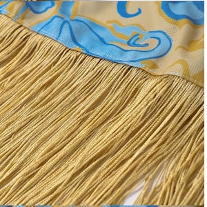 May include: Close-up of a decorative textile with a yellow fringe. The fabric above the fringe has a yellow background with blue and yellow swirling patterns. The fringe is made of many strands of yellow thread.