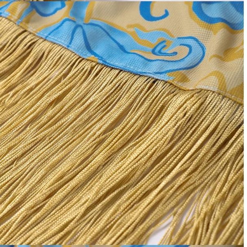 May include: Close-up of a decorative textile with a yellow fringe. The fabric above the fringe has a yellow background with blue and yellow swirling patterns. The fringe is made of many strands of yellow thread.