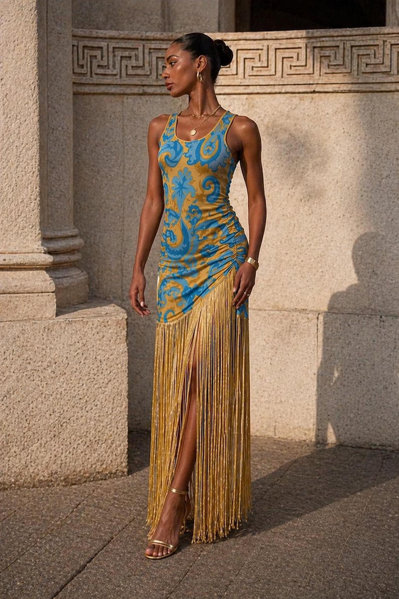 May include: A sleeveless, floor-length dress with a blue and gold paisley pattern. The dress features a high slit and is adorned with long, gold fringe. The model is wearing gold sandals and jewelry.