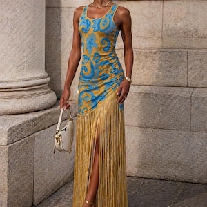 May include: A sleeveless, floor-length dress with a blue and gold paisley pattern. The dress features a high slit and a fringe detail at the hem. The model is wearing gold sandals and carrying a white handbag.