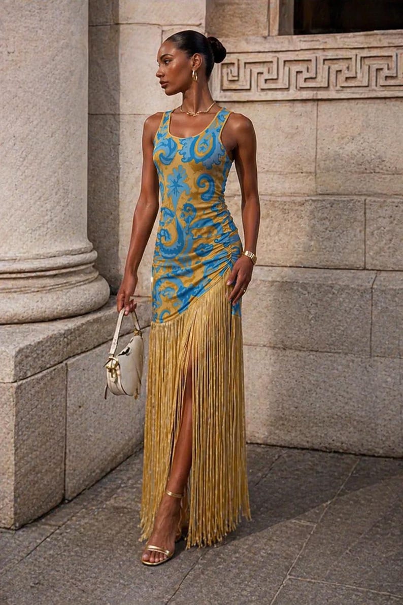 May include: A sleeveless, floor-length dress with a blue and gold paisley pattern. The dress features a high slit and a fringe detail at the hem. The model is wearing gold sandals and carrying a white handbag.