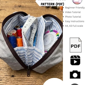 May include: A gray fabric organizer pouch with a brown zipper, featuring a light blue and white striped interior lining. The pouch contains colorful sewing thread spools, a white measuring tape, and a round pin cushion with multicolored pins.