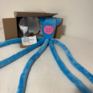 Puede incluir: Un peluche de pulpo azul con una gran cara de bot&oacute;n rosa y largos y suaves tent&aacute;culos. El juguete est&aacute; parcialmente dentro de una caja de cart&oacute;n abierta, con una nota manuscrita que dice: "Hey Janess, Look what I found in Gramma's Trunk. Remember? July 15".