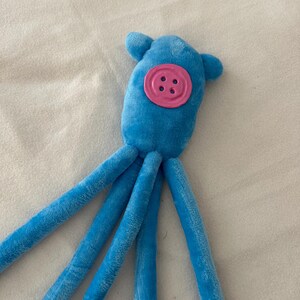 Puede incluir: Un juguete de pulpo de peluche azul con un detalle de bot&oacute;n rosa. El juguete tiene una cabeza redonda con dos peque&ntilde;as protuberancias en forma de oreja y m&uacute;ltiples tent&aacute;culos largos y suaves. El bot&oacute;n es de un color rosa m&aacute;s claro y tiene cuatro ojales.