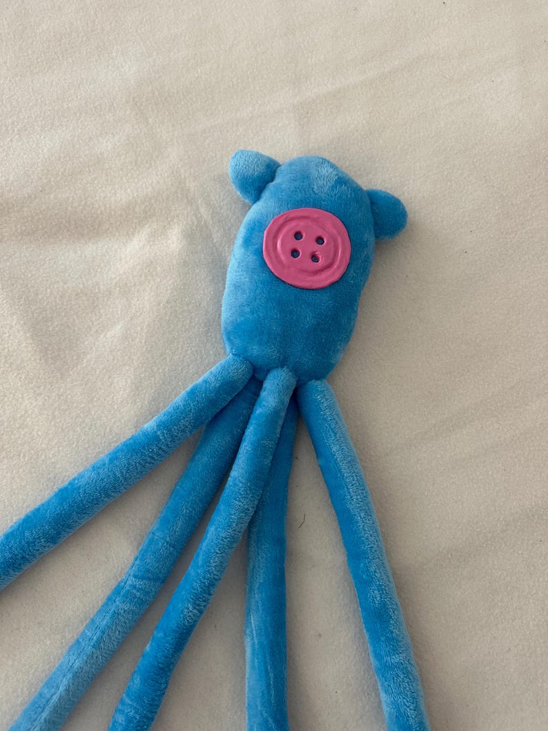 Puede incluir: Un juguete de pulpo de peluche azul con un detalle de bot&oacute;n rosa. El juguete tiene una cabeza redonda con dos peque&ntilde;as protuberancias en forma de oreja y m&uacute;ltiples tent&aacute;culos largos y suaves. El bot&oacute;n es de un color rosa m&aacute;s claro y tiene cuatro ojales.