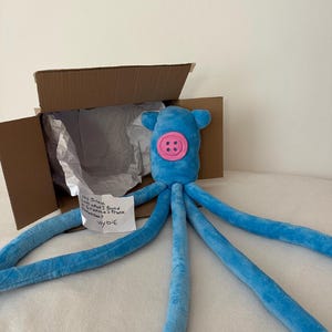 Puede incluir: Un juguete de peluche de pulpo azul con una cara de bot&oacute;n rosa y largos tent&aacute;culos. El juguete est&aacute; dentro de una caja de cart&oacute;n abierta con papel de seda. Una nota manuscrita dice: "Hey Janey, Look what I found in Grandma's trunk. Remember? Wybie."