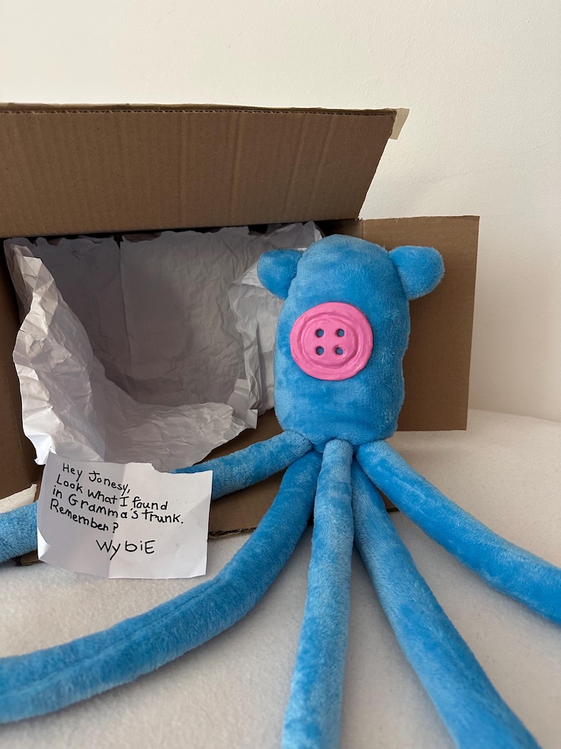 Puede incluir: Un juguete de peluche de pulpo azul con una cara de bot&oacute;n rosa y ocho largos tent&aacute;culos. El juguete est&aacute; dentro de una caja de cart&oacute;n abierta con papel de seda blanco. Una nota manuscrita dice: "Hey Jonesy, Look what I found in Gramma's trunk. Remember? Wybie."