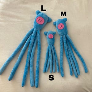 Puede incluir: Tres peluches azules de diferentes tama&ntilde;os, etiquetados L, M y S. Cada juguete tiene un bot&oacute;n rosa en la cara y m&uacute;ltiples ap&eacute;ndices largos y delgados. Los juguetes est&aacute;n colocados sobre una superficie clara.