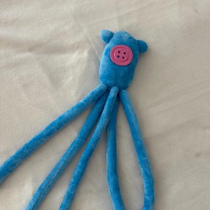 Puede incluir: Un juguete de peluche de pulpo azul con cuatro largos tent&aacute;culos. El juguete tiene un bot&oacute;n rosa para la cara y dos peque&ntilde;as orejas azules. El peluche es suave y parece estar hecho de un material difuso.