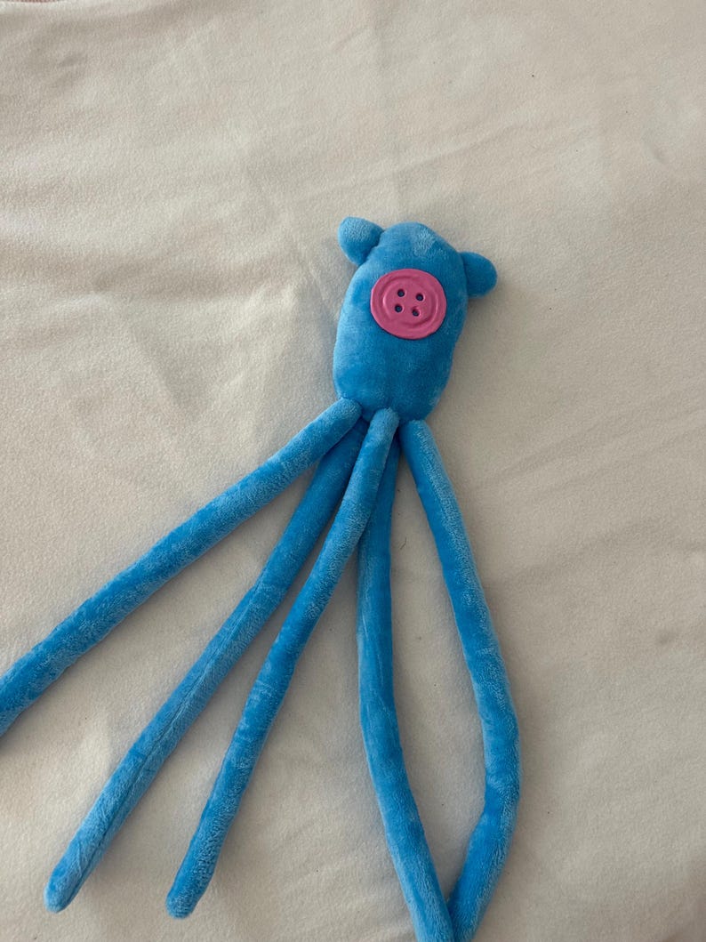Puede incluir: Un juguete de peluche de pulpo azul con cuatro largos tent&aacute;culos. El juguete tiene un bot&oacute;n rosa para la cara y dos peque&ntilde;as orejas azules. El peluche es suave y parece estar hecho de un material difuso.