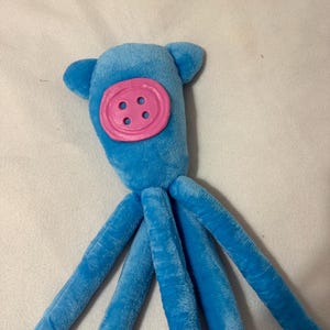 Puede incluir: Un juguete de pulpo azul de peluche con una cara de bot&oacute;n rosa y cuatro ojales. El juguete tiene dos peque&ntilde;os ap&eacute;ndices en forma de oreja en la parte superior de su cabeza y ocho largos tent&aacute;culos azules.