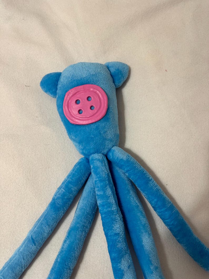 Puede incluir: Un juguete de pulpo azul de peluche con una cara de bot&oacute;n rosa y cuatro ojales. El juguete tiene dos peque&ntilde;os ap&eacute;ndices en forma de oreja en la parte superior de su cabeza y ocho largos tent&aacute;culos azules.