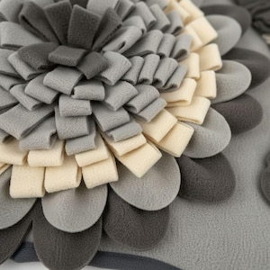 May include: A gray, cream, and dark gray felt flower-shaped pillow. The pillow features a layered design with petal-like shapes in varying shades of gray and cream, creating a textured, floral appearance. The pillow is a decorative home accent.