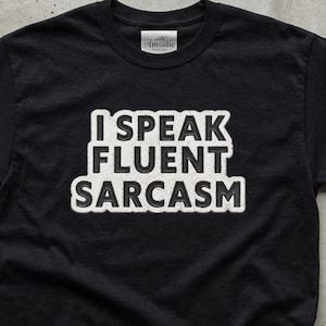 Pode incluir: T-shirt preta com o texto branco "I SPEAK FLUENT SARCASM". A camisa é exibida em uma superfície com uma xícara de café, um livro e óculos.