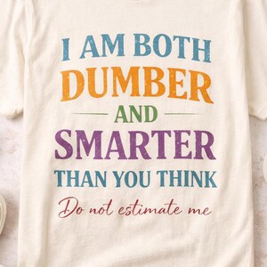 Op de afbeelding: Crèmekleurig T-shirt met de tekst "I AM BOTH DUMBER AND SMARTER THAN YOU THINK. Do not estimate me" in blauw, oranje, paars en rood. De afbeelding bevat witte sneakers en een kopje koffie.