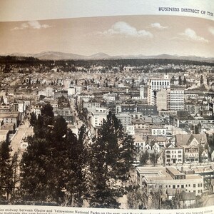 Op de afbeelding: Een zwart-wit luchtfoto van het zakendistrict van Spokane, Washington. De foto toont een dichtbevolkt stedelijk gebied met veel gebouwen en straten. De foto is gemaakt vanuit een hoog standpunt, waardoor een breed uitzicht op de stad wordt geboden.