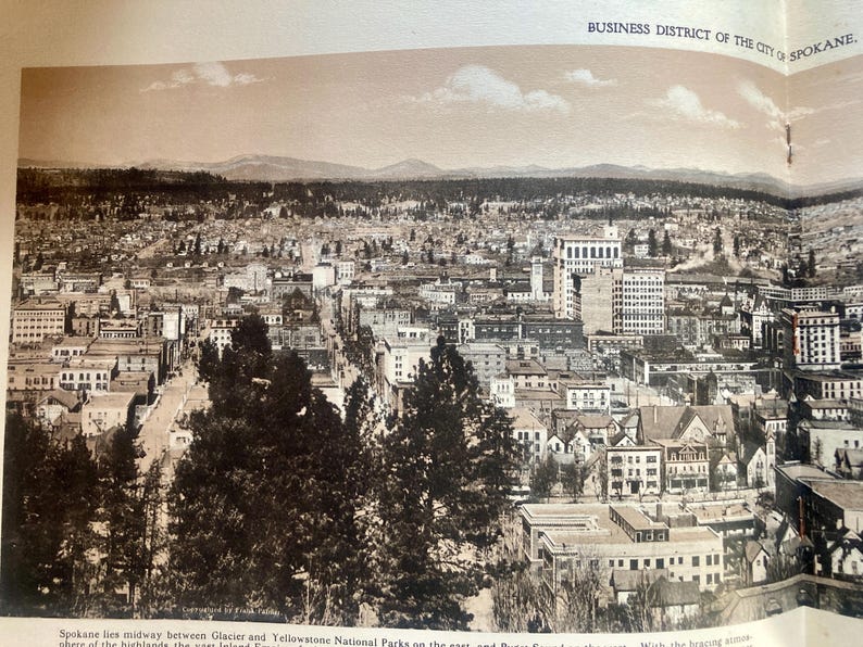 Op de afbeelding: Een zwart-wit luchtfoto van het zakendistrict van Spokane, Washington. De foto toont een dichtbevolkt stedelijk gebied met veel gebouwen en straten. De foto is gemaakt vanuit een hoog standpunt, waardoor een breed uitzicht op de stad wordt geboden.
