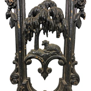 May include: A black cast iron architectural panel featuring a central relief of a willow tree and a resting lamb. The frame is decorated with floral rose motifs at the top corners and scrollwork details, showing signs of paint wear and chipping.