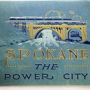 Op de afbeelding: Een vintage ansichtkaart met een gouden en blauwe illustratie van een brug met een trein die eroverheen rijdt. De tekst "Spokane The Power City" is gedrukt in gouden letters op een blauwe achtergrond.