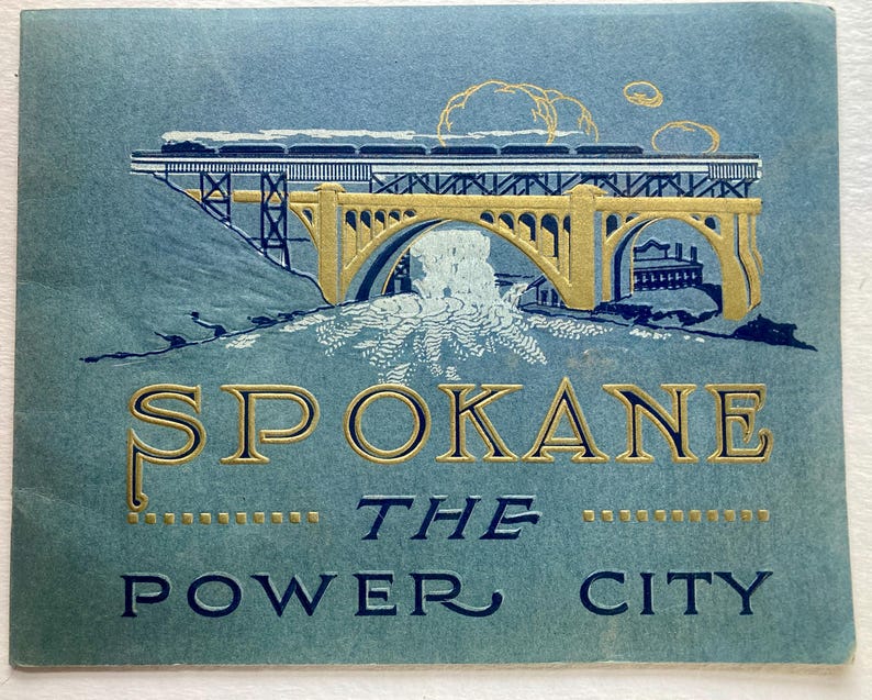 Op de afbeelding: Een vintage ansichtkaart met een gouden en blauwe illustratie van een brug met een trein die eroverheen rijdt. De tekst "Spokane The Power City" is gedrukt in gouden letters op een blauwe achtergrond.