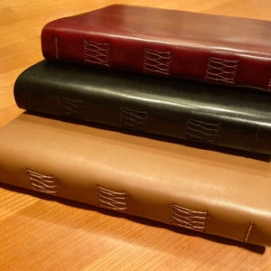 Puede incluir: Tres diarios encuadernados en cuero apilados uno encima del otro. El diario superior es burdeos, el diario del medio es negro y el diario inferior es marr&oacute;n. Los tres diarios tienen costuras decorativas a lo largo del lomo.