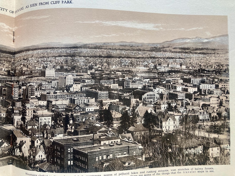 Op de afbeelding: Een zwart-wit luchtfoto van een stad. De stad is dichtbevolkt met gebouwen en bomen. De foto is genomen vanuit een hoog standpunt, waardoor een panoramisch uitzicht op de stad wordt geboden.