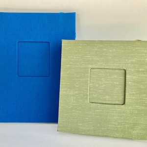 May include: Two photo albums, one blue and one green, with a rectangular window for a photo. Both albums have a textured fabric cover.