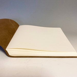 Puede incluir: Un diario encuadernado en cuero marr&oacute;n con p&aacute;ginas en blanco est&aacute; abierto para revelar el papel blanco en el interior. El diario est&aacute; acostado sobre una superficie blanca.