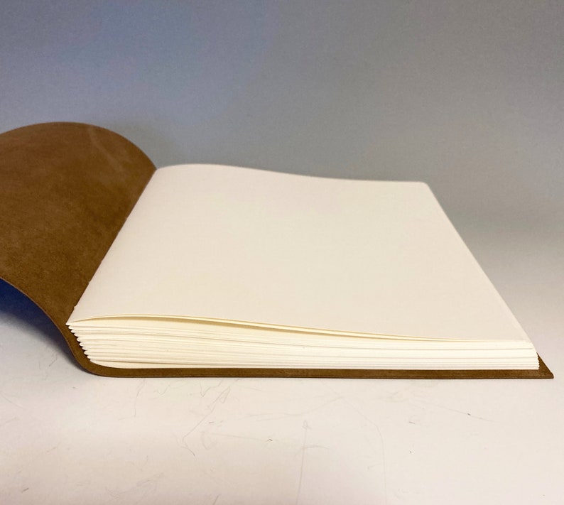 Puede incluir: Un diario encuadernado en cuero marr&oacute;n con p&aacute;ginas en blanco est&aacute; abierto para revelar el papel blanco en el interior. El diario est&aacute; acostado sobre una superficie blanca.