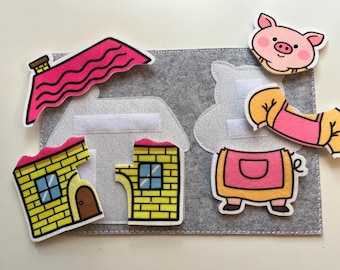 Puede incluir: Un tablero de fieltro con piezas para un cuento infantil. Incluye una casa con tejado rosa, casas de ladrillo amarillas, un cerdo y un atuendo de cerdo. El fondo es un tablero de fieltro gris con velcro blanco.