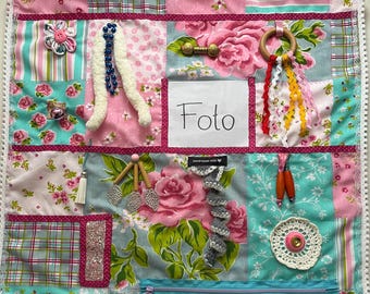 Puede incluir: Una manta de actividades sensoriales cuadrada con un patchwork de telas florales y de cuadros en rosa, turquesa y blanco. Incluye varios elementos táctiles como cintas, cuentas, un anillo de madera, una cremallera y un cuadrado blanco central con la etiqueta Foto.