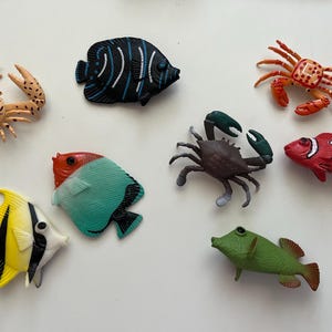 Puede incluir: Una colecci&oacute;n de juguetes de criaturas marinas coloridas, incluyendo cangrejos y varios peces. Los peces son en tonos de amarillo, negro, rojo, blanco, verde y azul. Los cangrejos son de color beige, marr&oacute;n y naranja. Los juguetes est&aacute;n dispuestos sobre una superficie blanca.