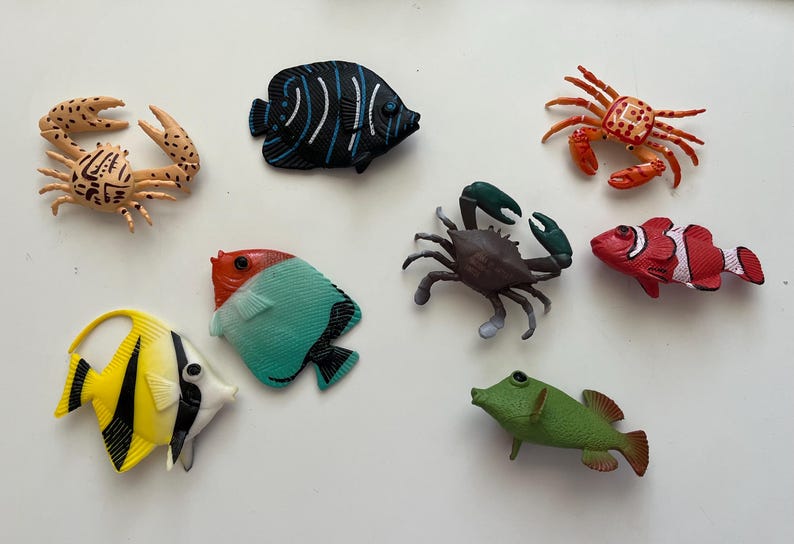 Puede incluir: Una colecci&oacute;n de juguetes de criaturas marinas coloridas, incluyendo cangrejos y varios peces. Los peces son en tonos de amarillo, negro, rojo, blanco, verde y azul. Los cangrejos son de color beige, marr&oacute;n y naranja. Los juguetes est&aacute;n dispuestos sobre una superficie blanca.