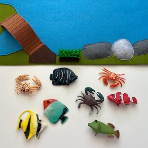 Puede incluir: Un tapete de juego de fieltro con un estanque azul, un puente marr&oacute;n y hierba verde. Varias criaturas marinas de juguete, incluyendo cangrejos y peces coloridos, est&aacute;n dispuestas alrededor del tapete, creando una escena submarina.