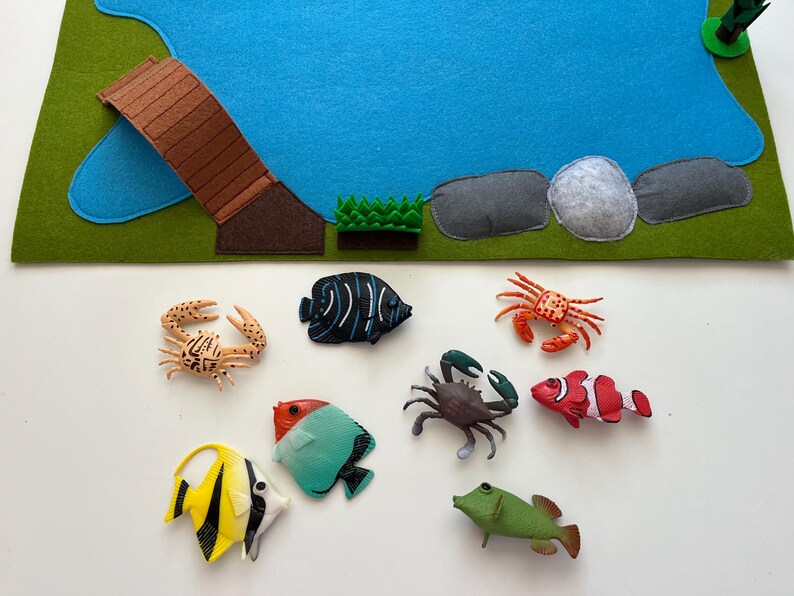 Puede incluir: Un tapete de juego de fieltro con un estanque azul, un puente marr&oacute;n y hierba verde. Varias criaturas marinas de juguete, incluyendo cangrejos y peces coloridos, est&aacute;n dispuestas alrededor del tapete, creando una escena submarina.