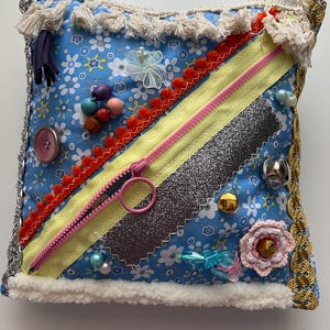 Könnte beinhalten: Ein quadratisches sensorisches Kissen mit blauem Blumenmuster, verziert mit verschiedenen Texturen und Verzierungen. Es verfügt über einen rosa Reißverschluss, Glitzerstoff, bunte Perlen und dekorative Verzierungen. Die Kanten sind mit weißem Kunstfell und Quasten versehen.